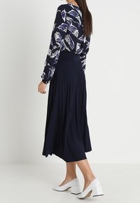 Abito blu navy con motivo floreale, gonna plissettata, maniche lunghe e vita affusolata. Abbinato a scarpe bianche con tacco quadrato.
