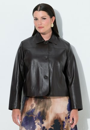 Frau mit langen dunklen Haaren, die goldene Ohrringe, eine schwarze Lederjacke mit Knöpfen und einen Rock mit Blumenmuster vor weißem Hintergrund trägt.