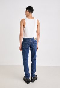 Biela bezrukávová blúzka a modré džínsy s charakteristickými zadnými vreckami. Čierne masívne topánky dopĺňajú outfit. Minimalistické pozadie.