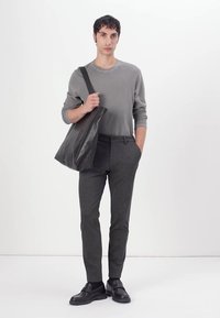 Graues langärmeliges Hemd, dunkelgraue schmal geschnittene Hose und schwarze Schuhe. Hält eine schwarze Ledertasche und steht vor einem weißen Hintergrund.