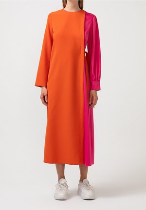 Frau steht und trägt ein langärmeliges Midikleid mit einfarbig orangefarbenem Vorderteil und leuchtend pinkem Seitenpanel sowie weißen Sneakers vor weißem Hintergrund.