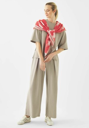 Frau trägt beige Hosen mit weitem Bein und ein übergroßes T-Shirt, mit einem rot gemusterten Schal, der um ihre Schultern gebunden ist, und steht vor weißem Hintergrund.