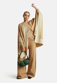 Beżowo-biała, oversize'owa narzutka z kanciastym wzorem, noszona na beżowym swetrze i spodniach. Zielona mini torebka z kieszonkami, czarne obuwie.
