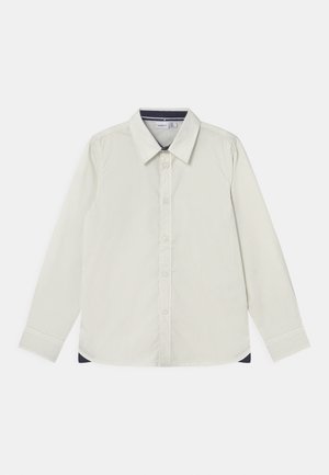 Camisa de manga larga color beige claro con botones, cuello puntiagudo, tela texturizada y pequeños detalles oscuros en el interior del cuello y los laterales del dobladillo.