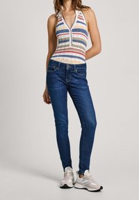 Kvinna iklädd en ärmlös, flerfärgad randig topp, mörkblå slim jeans och vita sneakers, stående framför en enkel bakgrund.