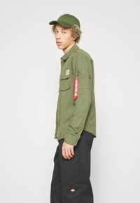 Olive green button-up shirt with two chest pockets and a side zip pocket, featuring a red label. Worn with black pants and a matching cap.