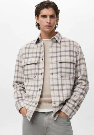 Camisa de franela a cuadros en gris y blanco con dos bolsillos en el pecho, botón frontal y un corte relajado, superpuesta sobre un suéter beige.
