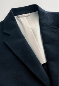 Blazer in lana blu con colletto a scollo e foderato in crema, con una texture liscia e un sottile dettaglio di cucitura vicino al risvolto.