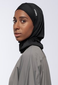Donna che indossa un hijab nero con il testo "kamak" e una maglietta grigia ampia, rivolta di lato contro uno sfondo chiaro e semplice.