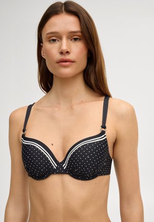 Vrouw met lang bruin haar, dragend een zwarte beha met witte polka dots en dubbele witte strepen op de bandjes, tegen een effen lichte achtergrond.