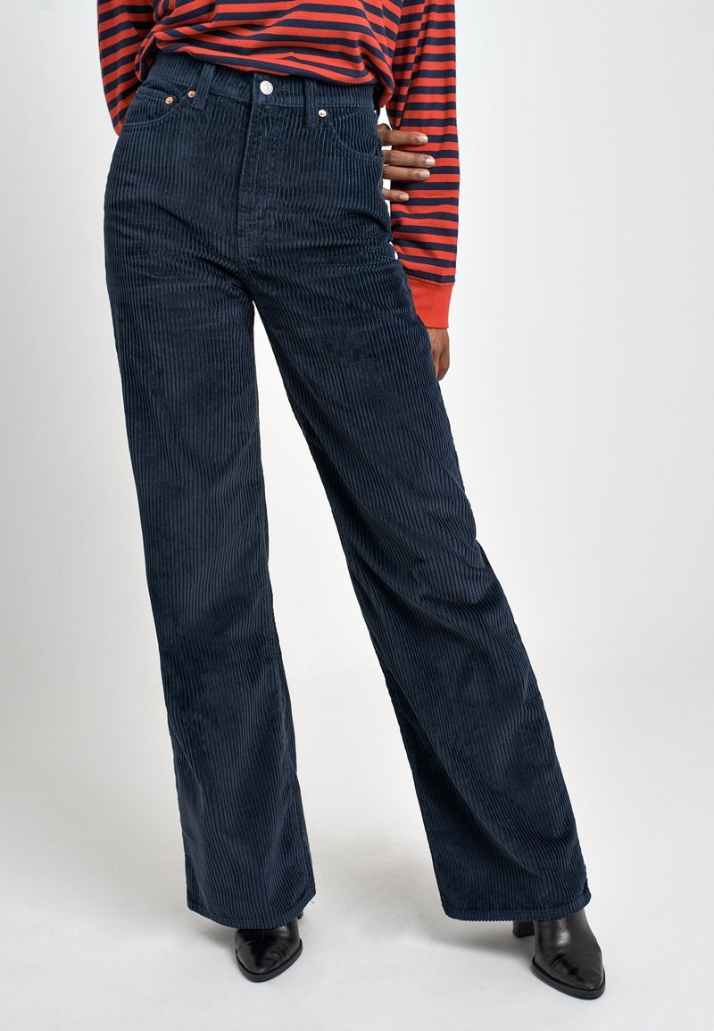 Femme portant un pantalon en velours côtelé bleu marine taille haute à jambes larges, un haut à manches longues rayé rouge et bleu marine, et des bottines noires à talons.