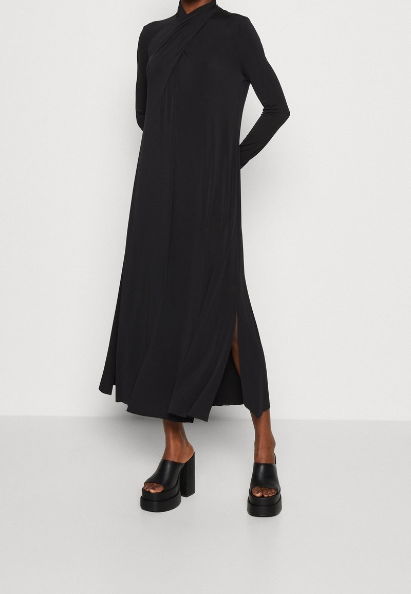 Musta, pitkähihainen mekko, jossa on laskostettu muotoilu ja sivuaukko, valmistettu sileästä kankaasta. Yhdistettynä paksupohjaisiin mustiin korkokenkiin.