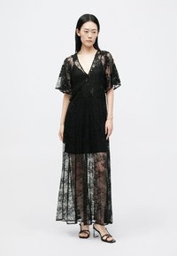 Juoda nėrinių maksi suknelė su trumpomis plazdančiomis rankovėmis, V formos iškirpte ir skaidriu sluoksniu. Apima sudėtingus gėlių raštus ir laisvą siluetą.