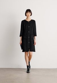 Abito nero con maniche a tre quarti, tessuto texturizzato, tasche anteriori e chiusura con bottoni. Indossato con stivali alla caviglia e una pochette.