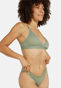Zelená bavlněná podprsenka s V-švihem a tenkými ramínky, v kombinaci se zelenými bikini kalhotkami, které mají široký pás a minimální pokrytí.