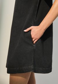 Schwarzes Jeanskleid mit einer Seitentasche, sichtbaren Nähten und einem knielangen Saum. Der Stoff hat eine glatte Textur mit einem leichten Glanz.