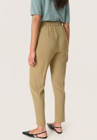 Beige elastīgas jostas bikses ar sašaurinātu piegriezumu, izgatavotas no mīksta auduma. Apvienotas ar melniem slingback apaviem. Redzami šuvju detaļas aizmugurē.