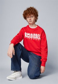 Felpa rossa con logo bianco "DIESEL", abbinata a jeans blu larghi e sneakers di colore chiaro. Sfondo neutro, soggetto in ginocchio.