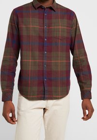 Camicia in flanella a quadri nei colori verde scuro, burgundy e blu navy, con chiusura a bottoni, maniche lunghe e una tasca sul petto.