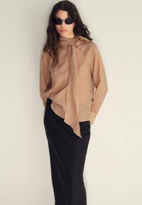Hellbeige Bluse mit gebundenem Kragen, kombiniert mit einem schwarzen, eng anliegendem Maxirock. Modell trägt dunkle Sonnenbrille, Haare wellig.