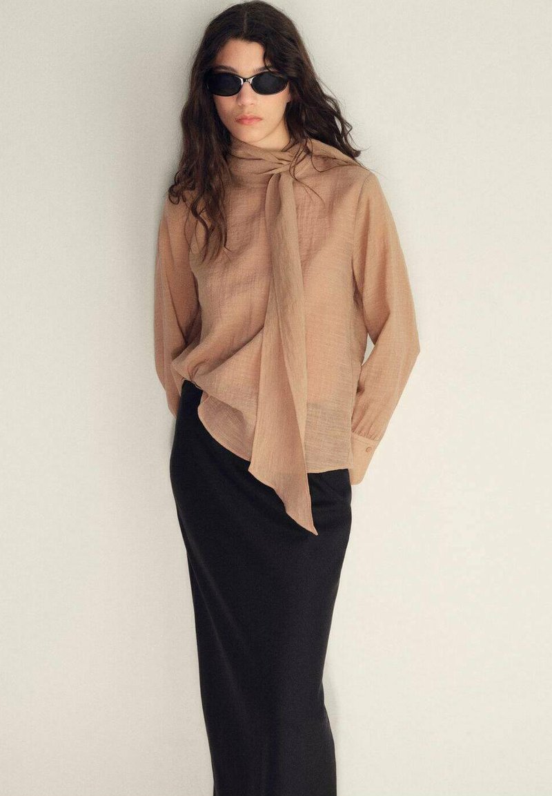 Ljus beige blus med knytdetalj vid halsen, kombinerad med en svart, åtsittande maxikjol. Modellen bär mörka solglasögon och har vågigt hår.