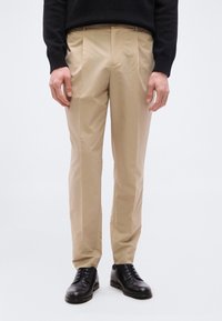 Άτομο που φοράει μπεζ ραμμένο παντελόνι, μαύρα επίσημα παπούτσια και μαύρο μακρυμάνικο τοπ, στέκεται μπροστά σε απλό λευκό φόντο.