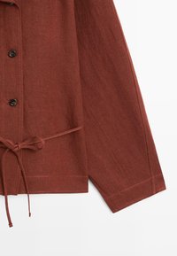 Lang shirt met lange mouwen in roestkleur, met zwarte knopen en een taillekoord, op een witte achtergrond.