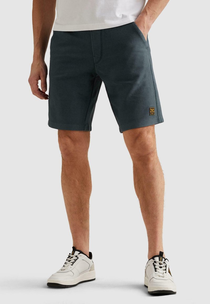Homme portant un short décontracté gris foncé et des baskets blanches, debout avec une main dans la poche sur fond uni.