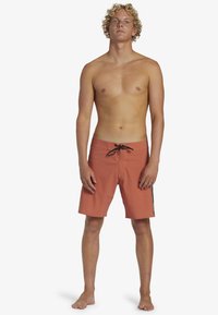 Shorts de bain rouge-orangé avec un cordon noir, coupe mi-genou, devant plat et petit logo sur le côté. Portés par un mannequin masculin debout.