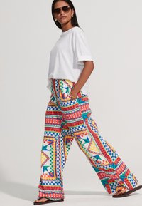 Pantalon à jambes larges avec motifs géométriques colorés en rouge, bleu, vert et jaune, associé à un t-shirt blanc et des lunettes de soleil.