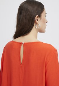 Rote Bluse mit strukturierter Oberfläche, ausgestattet mit einem Schlüssellochverschluss im Rücken und einer lockeren Passform. Einfaches Design mit langen Ärmeln.