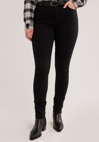 Naine, kes kannab musti kitsaid teksapükse musta vööd, musti kontsakingi ja mustvalget ruudulist särki.