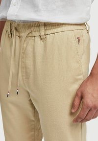 Beige trekbroek met een elastische tailleband, zijzakken en een geborduurd ontwerp op de heup. Glad, lichtgewicht stof met een gestructureerde afwerking.