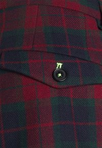 Rood en groen geruit stof met een gestructureerde weving, met een zwarte knop en een klein, felgroen logo geborduurd op de zak.