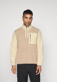 Fleece-pullover i beige og krem, med halvmeter glidelås, brystlomme med glidelås, og logo på venstre side. Myk tekstur, tettsittende design.