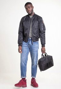 Zwart bomberjack, gestreept shirt, lichtblauwe jeans, rode sneakers, een zwarte tas vasthoudend. Voorzien van geribbelde manchetten en een zak met rits.