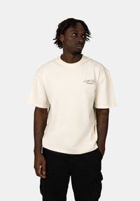 Cream-colored cotton t-shirt with a relaxed fit, short sleeves, and a small branded text detail on the chest. Worn with black cargo shorts.