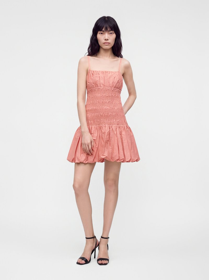 Jeune femme aux longs cheveux foncés portant une robe corail sans manches froncée et des sandales noires à talons hauts, debout devant un fond blanc uni.