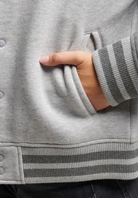 Sweat-shirt gris avec un tissu texturé, des poignets côtelés à rayures gris foncé, des boutons-pression et une poche latérale avec une main visible.