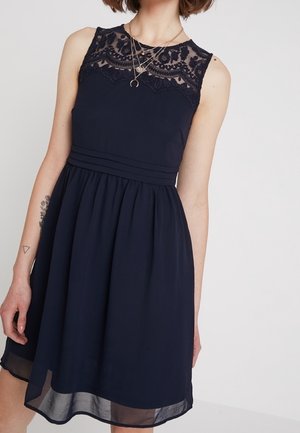 Navy blue dress with a lace neckline, sleeveless design, gathered waist, and a flowing skirt with a sheer overlay at the hem.