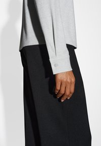 Maglione a costine grigio chiaro con maniche lunghe, caratterizzato da un dettaglio con bottone sul polsino, abbinato a pantaloni neri. Tessuto liscio e vestibilità rilassata.