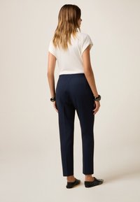 Donkerblauwe tapse broeken met een elastische tailleband, gecombineerd met een witte top met korte mouwen. Kenmerkt zich door zwarte platte schoenen met studs.