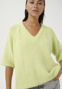 Pulover tricotat verde deschis, cu mânecă scurtă și decolteu în V. Textură moale, cu o croială lejeră și un aspect subtil pufos. Fără modele sau detalii vizibile.