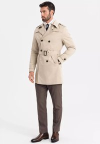 Beżowy płaszcz trench z podwójnym zapięciem, czarnymi guzikami i paskiem. Nosi się go na białej koszuli, zestawiony z brązowymi spodniami i skórzanymi butami.