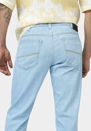 Hellblaue Jeans aus Denim, getragen von einer Person mit tätowiertem Arm, kombiniert mit einem hellgelben gemusterten Hemd, von hinten betrachtet.