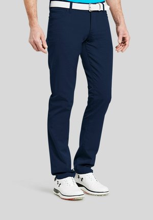 Man draagt marineblauwe broek, witte geweven riem en witte Under Armour golfschoenen, staand op een grijze achtergrond.