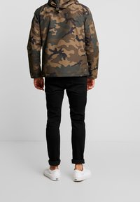 Chaqueta de camuflaje en tonos de verde y marrón, combinada con jeans negros y zapatillas blancas, vista desde la parte de atrás.