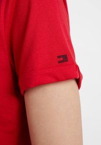 Acercamiento a una manga enrollada de una camisa roja con un pequeño logotipo negro en el puño. La tela parece suave y casual.