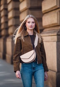 Mladá žena s dlouhými blond vlasy prochází kolem kamenné budovy, má na sobě hnědou bundu, modré džíny a krémovou crossbody tašku s širokým popruhem.