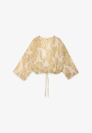 Mujer que lleva pantalones de pierna ancha en beige y verde oliva y una chaqueta ligera estampada con cordón, de pie contra un fondo liso y claro.